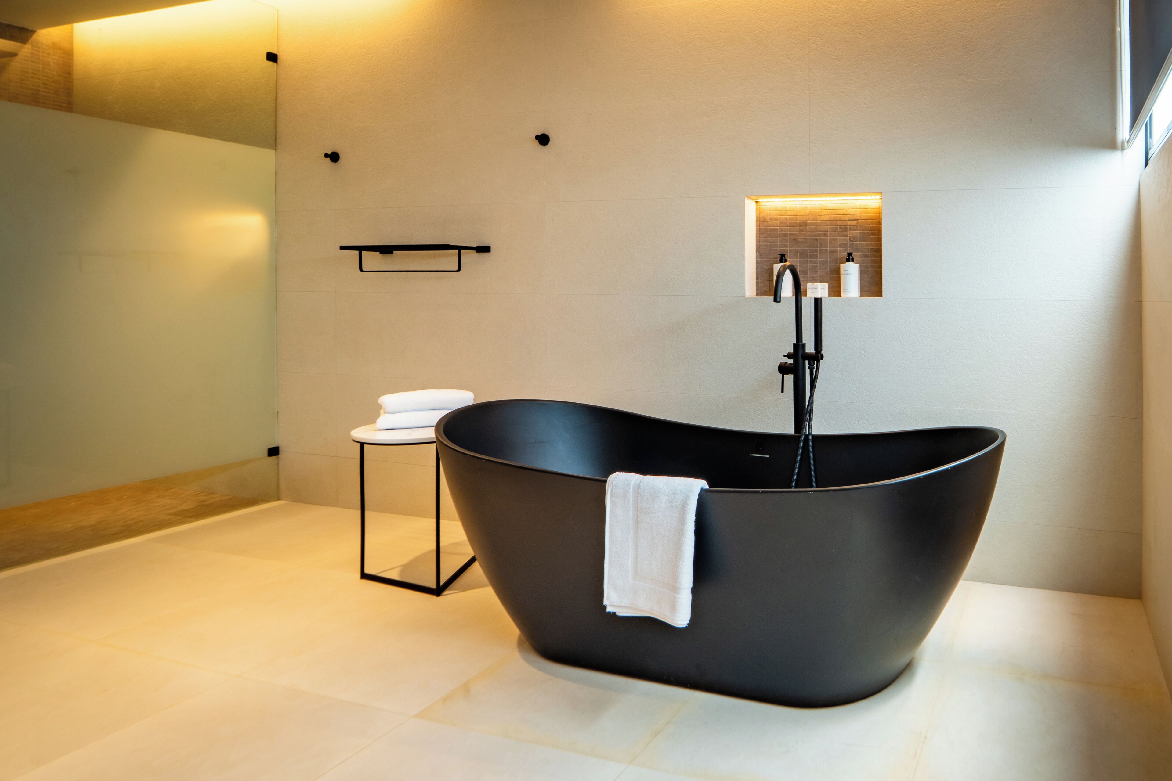 A modern bathroom with a freestanding bathtub, a small stool with a towel, and a wall-mounted shelf with toiletries.