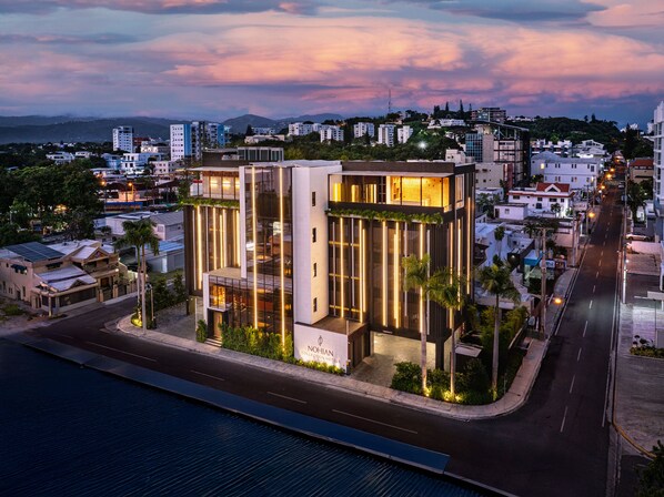 A modern building with a glass facade and vertical lighting accents, located in a cityscape with a view of the sea and mountains in the background. - Nohian Collection Hotels (Santiago de los Caballeros)