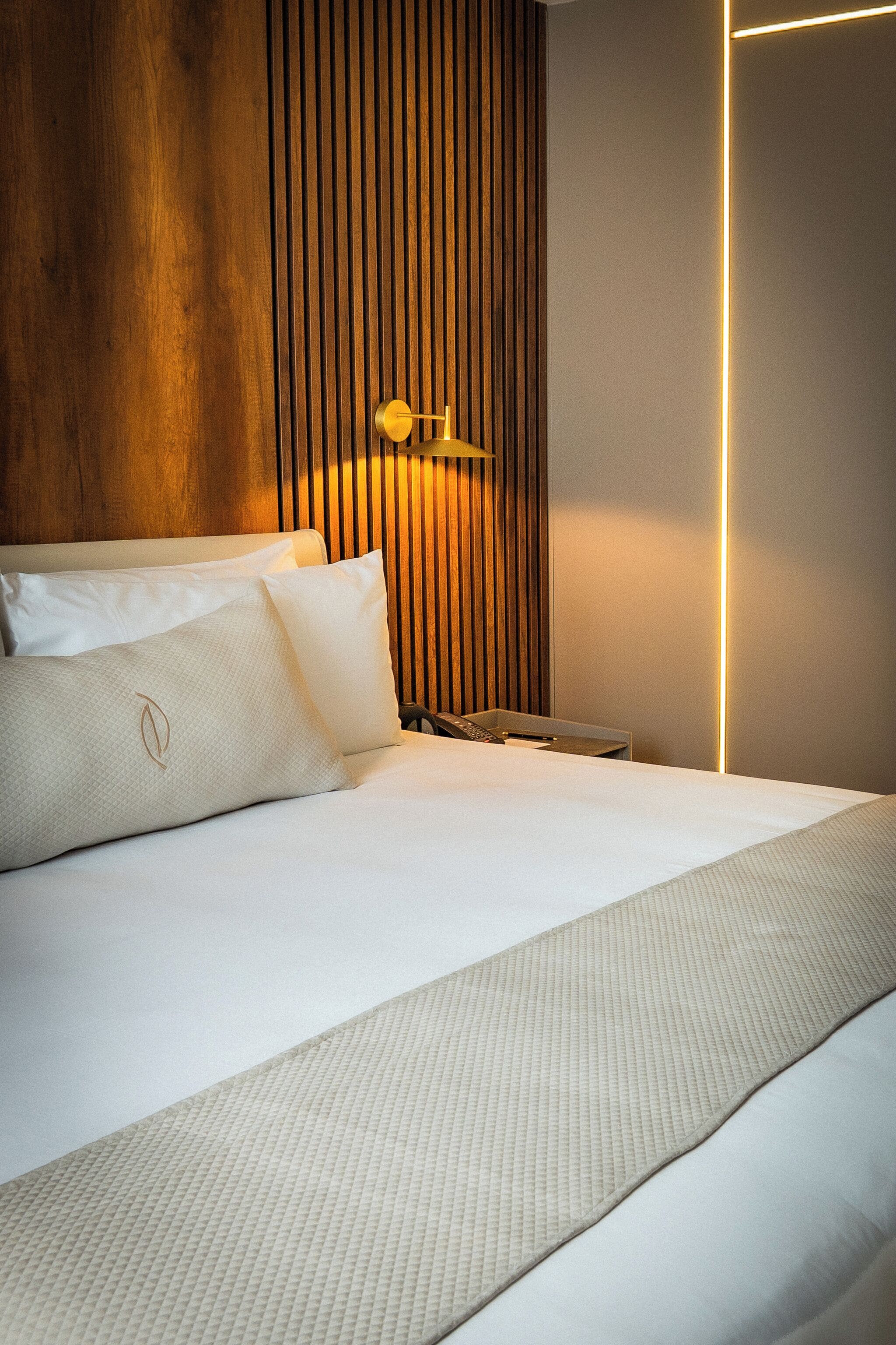 Ein ordentlich bezogenes Bett mit weißen Leinen und einer strukturierten Decke, ein Lattenrost aus Holz sowie eine Wandleuchte.