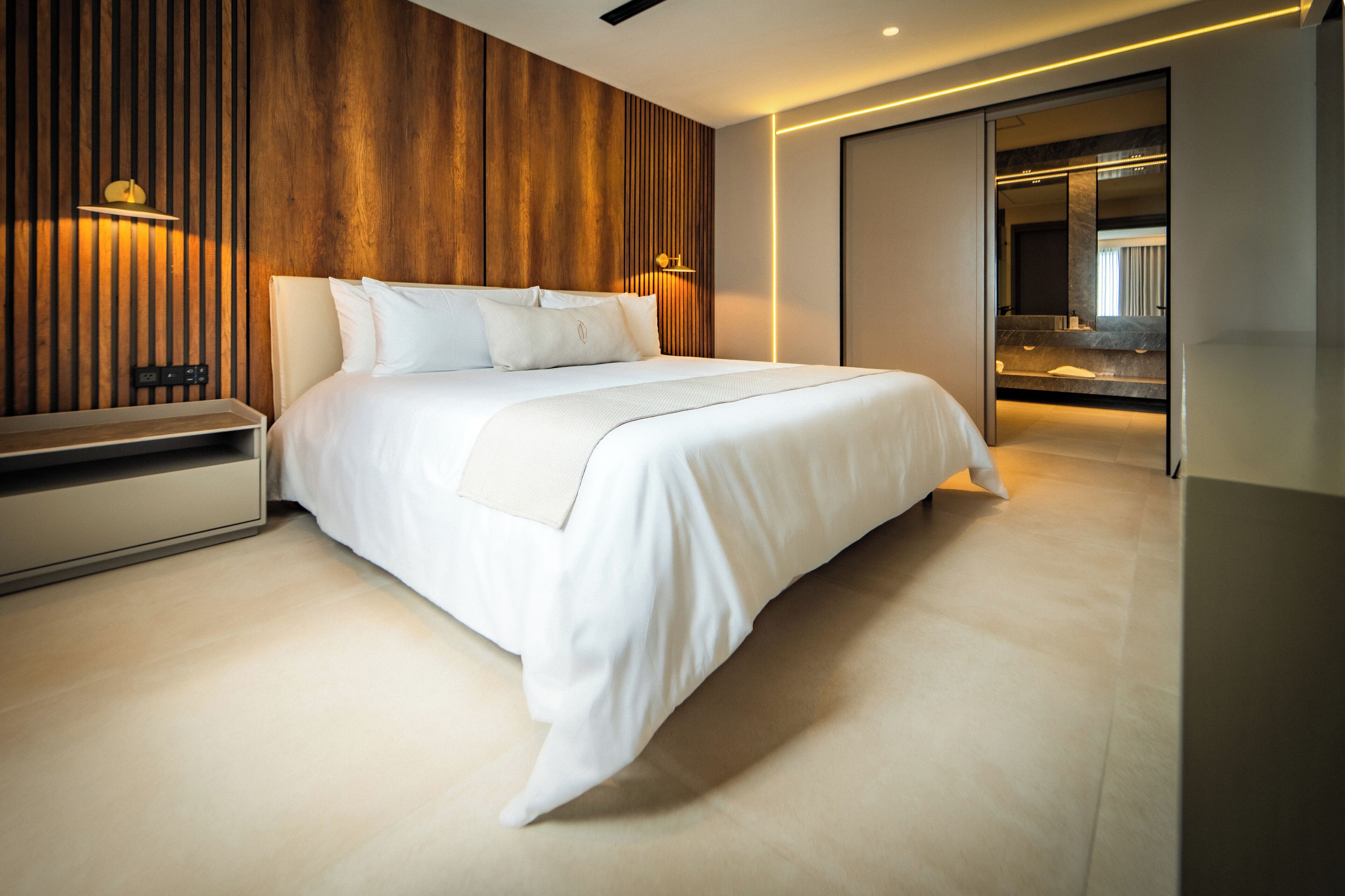 A modern hotel room with a large bed, wooden panel wall, and a bathroom visible through a sliding door.