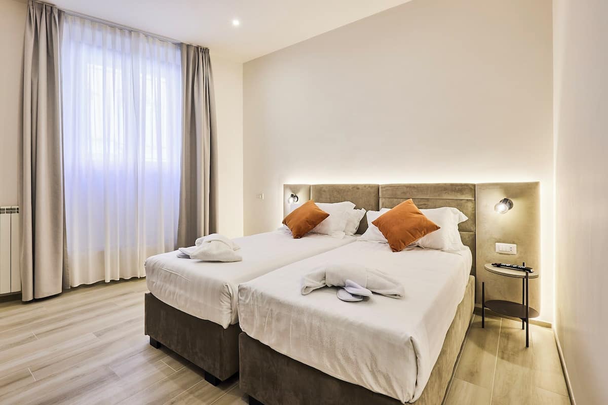 Habitación de hotel con dos camas, una mesita de noche y un ventanal con cortinas.