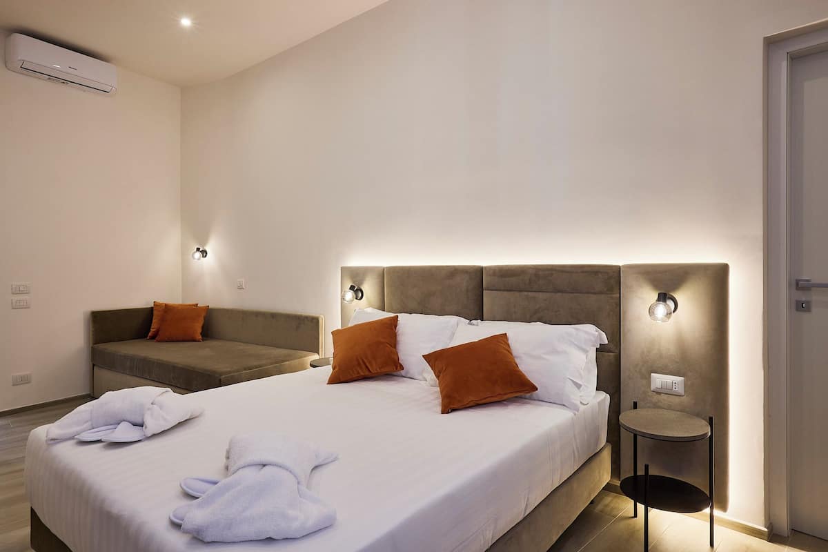 Habitación de hotel con una cama grande, dos mesitas de noche, un sofá y un aire acondicionado instalado en la pared.