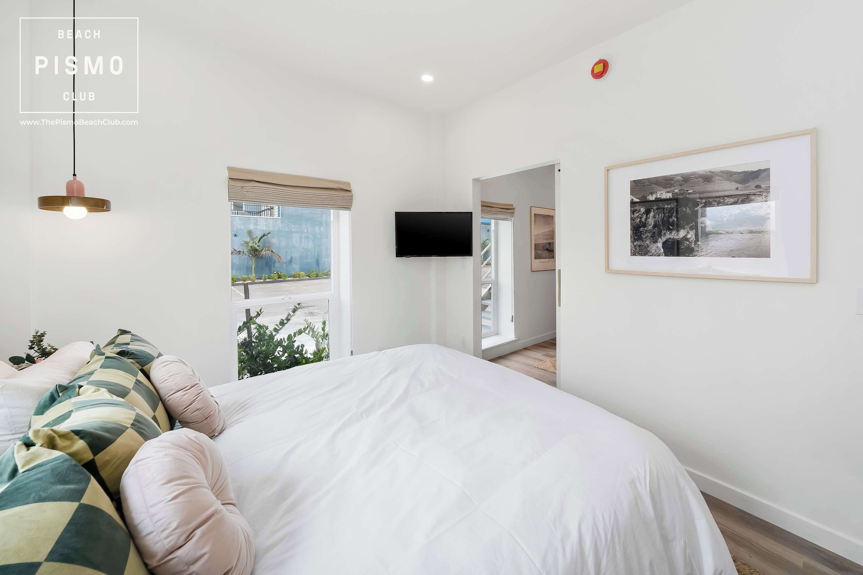 Un dormitorio con una cama grande, televisión, un cuadro enmarcado y una ventana con vistas a palmeras.