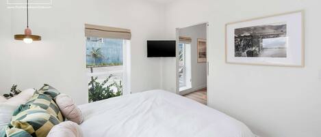 Un dormitorio con una cama grande, televisión, un cuadro enmarcado y una ventana con vistas a palmeras.