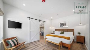 A bedroom with a bed, a chair, a TV, a door, and a sign for Beach Pismo Club. - Deluxe Downtown View Suite at Pismo Beach Club (Pismo Beach)