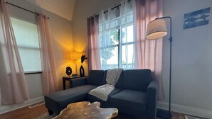 Ein Wohnzimmer mit einem dunkelgrauen Sofa, einem Holztisch, einer Stehlampe und einem Fenster mit durchscheinenden Vorhängen.