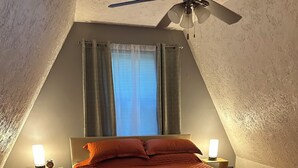 Ein Schlafzimmer mit einem Bett, zwei Nachttischen mit Lampen, einem Deckenventilator, einem Fenster mit Vorhängen und Holzböden.