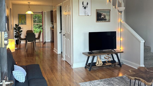 Ein geräumiger Wohnbereich mit hohen Decken, Holzboden, einer schwarzen Couch, einem Fernseher auf einem Holzständer und einer Treppe mit weißer Geländer.