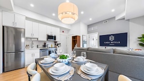 Una cocina moderna con electrodomésticos de acero inoxidable, un comedor con mesa preparada para cuatro personas, y una sala de estar con sofá gris.