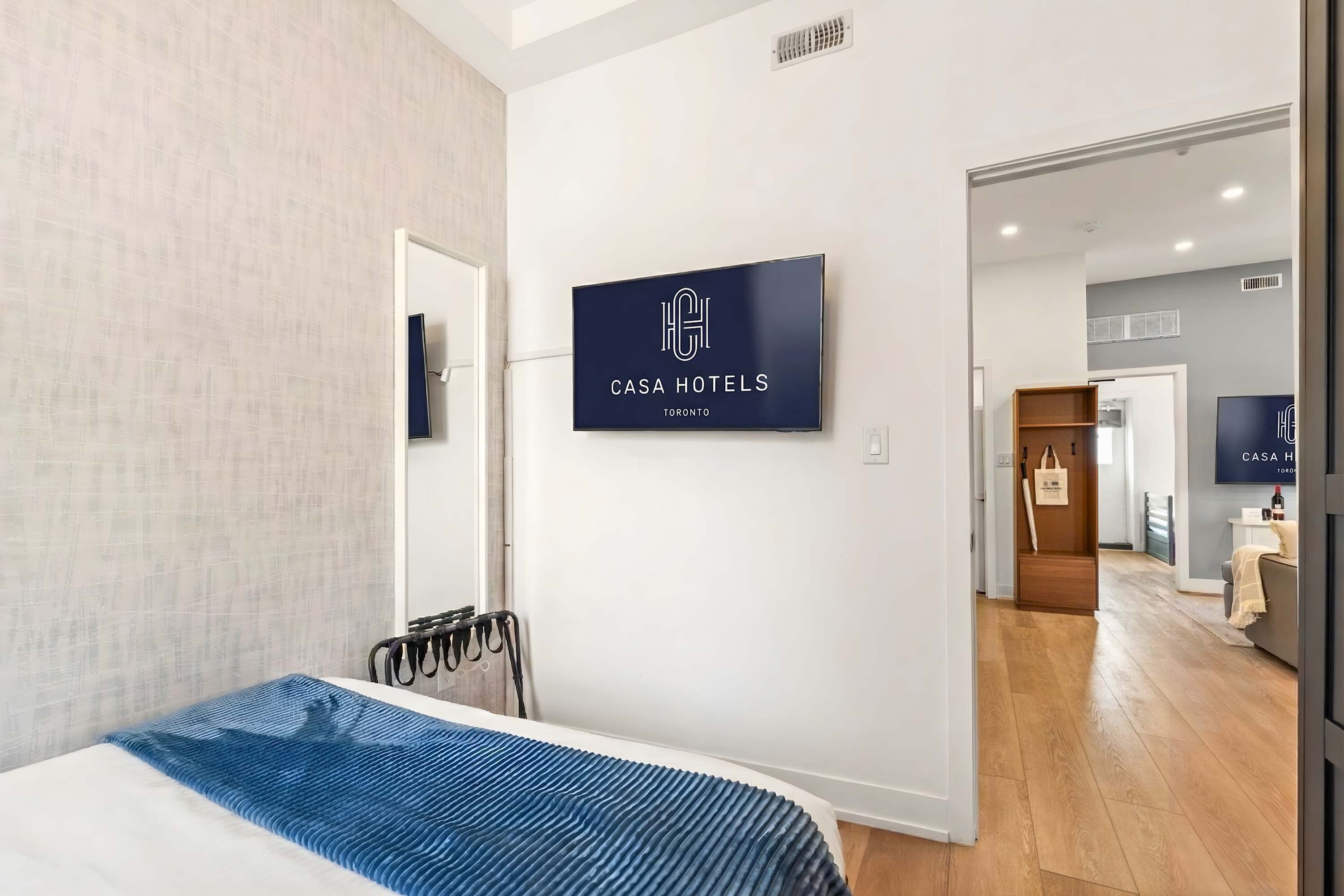 A modern hotel room with a bed, a TV mounted on the wall, and a visible hallway leading to other rooms.