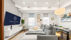 Una habitación de hotel moderna con cama, sofá, mesa de comedor y un televisor mostrando el nombre del hotel.