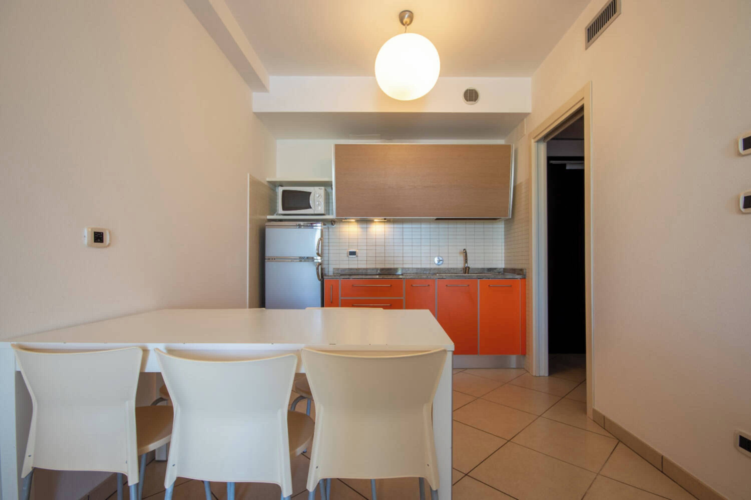 A compact kitchen with a dining area, featuring a white table and chairs, a microwave, a refrigerator, and orange cabinets.