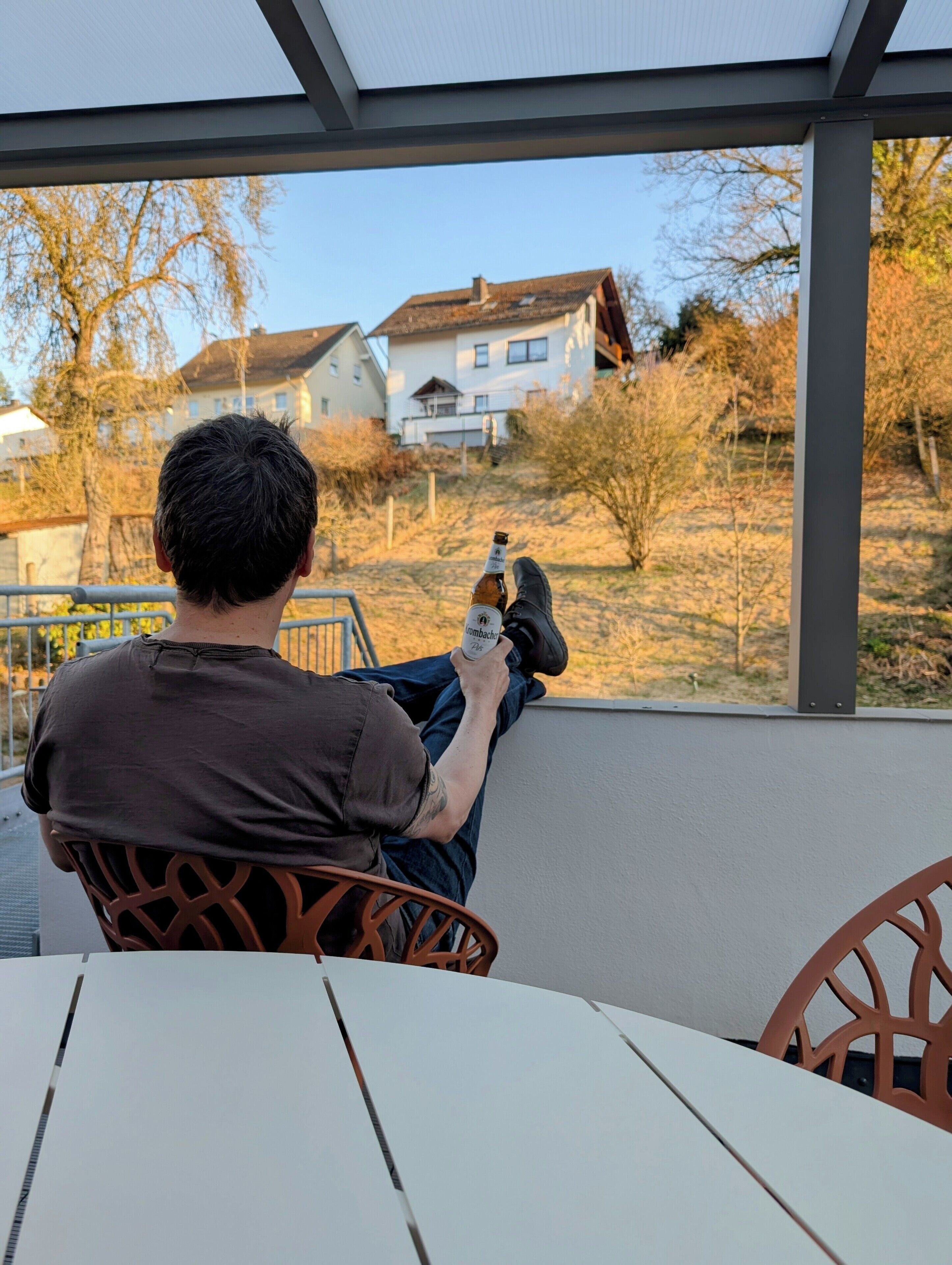 A person sitting at an outdoor table with a bottle of beer, overlooking a residential area with houses and trees.