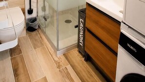 Un baño moderno con ducha de vidrio, inodoro blanco, tocador de madera y lavadora integrada.