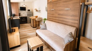 Un espacio habitable compacto con sofá, una mesita, una zona de cocina con microondas y refrigerador, y un televisor montado en la pared.