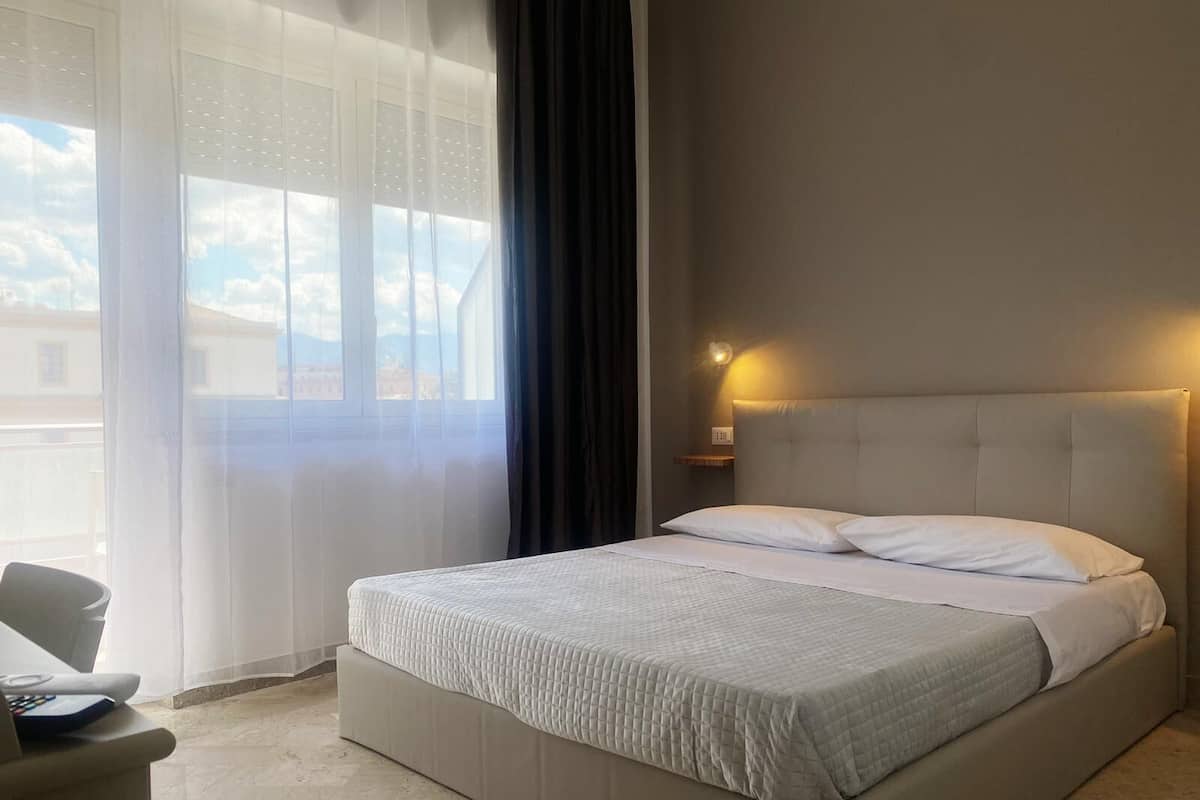 Ein Hotelzimmer mit einem großen Bett, einem Sessel, einem Nachttisch und einem Fenster mit durchsichtigen Vorhängen.