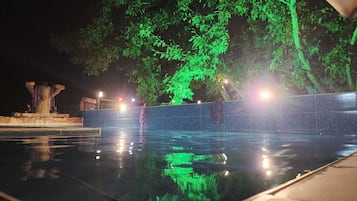 A nighttime swimming pool with a fountain, surrounded by trees and illuminated by green lights.
