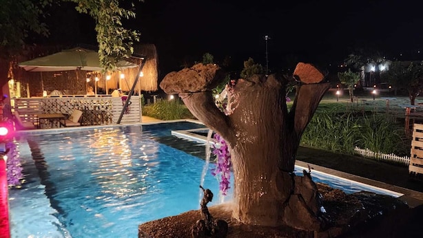 Zona de piscina nocturna con una fuente en forma de árbol y un área de estar con mesas y sillas.