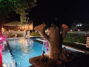 A nighttime pool area with a tree-shaped water feature and a seating area with tables and chairs.