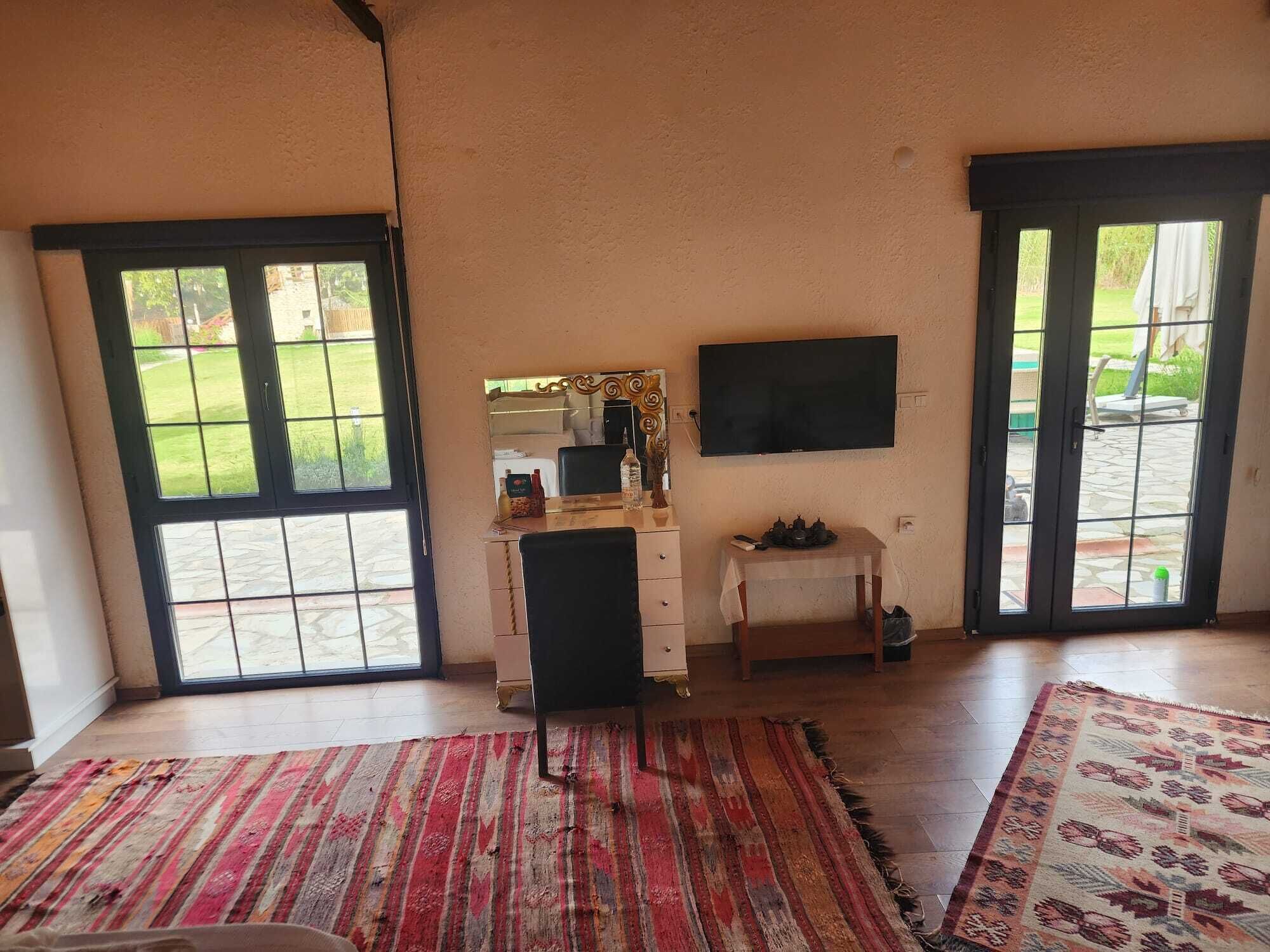 Een kamer met een groot raam, een televisie, een klein tafeltje en een kamerkleed met een patroon.