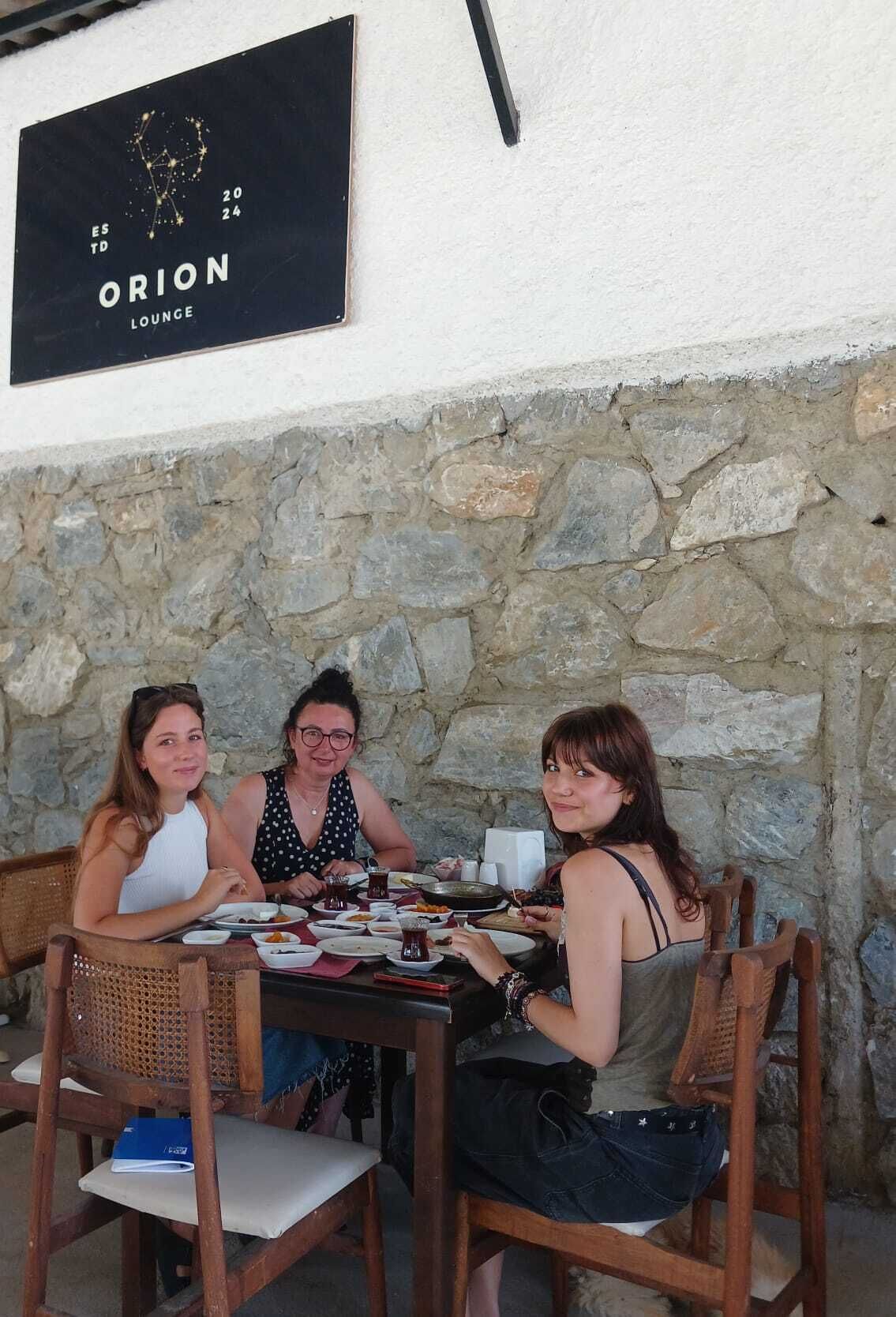 Tre personer sitter vid ett bord med en vägg av sten som bakgrund. Ovanför bordet hänger en skylt med texten: "ORION LOUNGE, grundat 2024".
