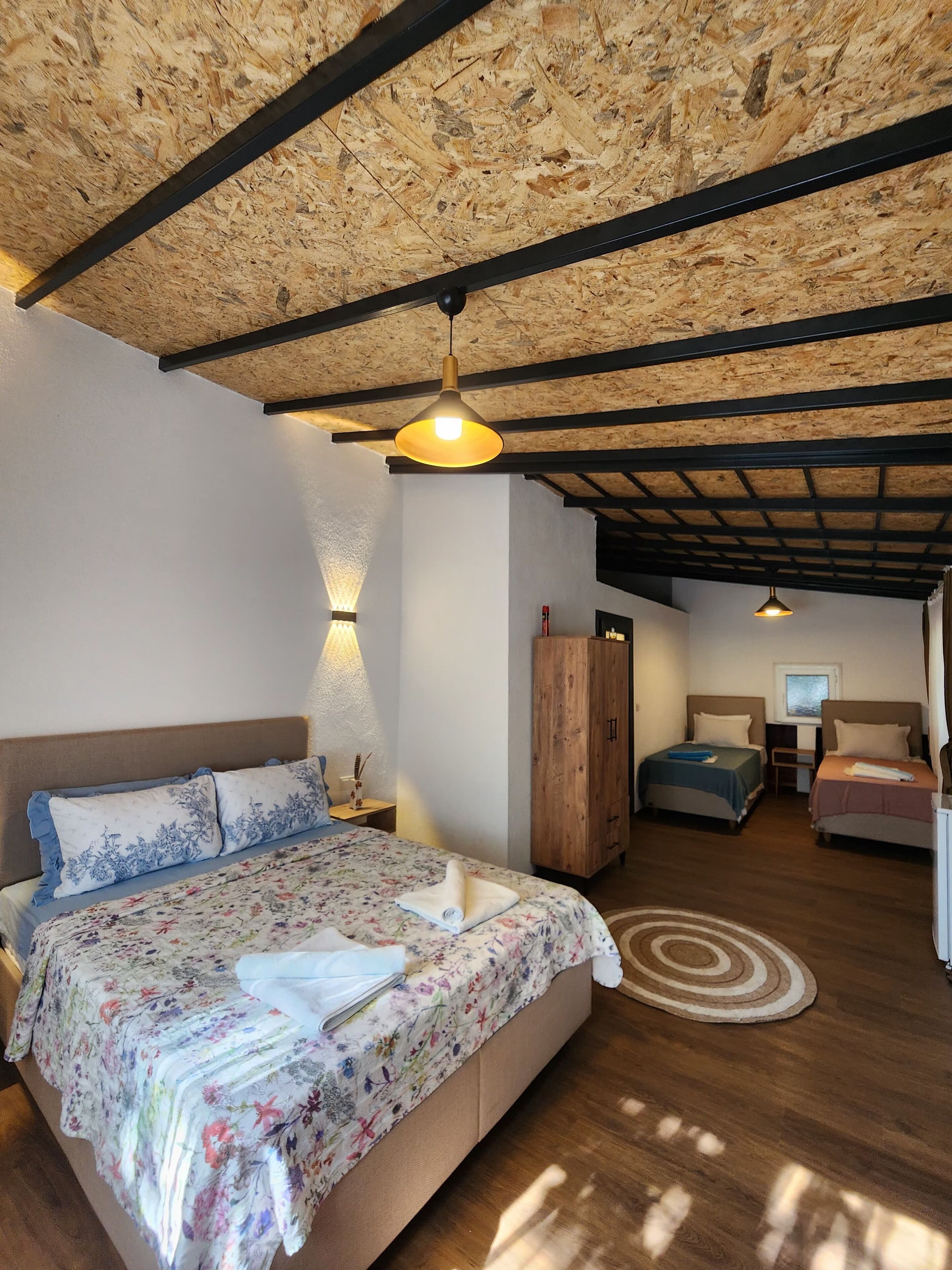 A bedroom with a wooden ceiling, a large bed with floral bedding, a wooden wardrobe, and a small desk with a chair.