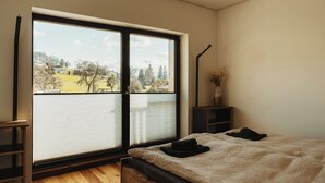 Une chambre avec une grande fenêtre, un lit, une table de chevet et un petit meuble.