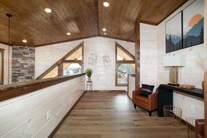 A spacious room with wooden floors, a stone accent wall, a large window, and a seating area with a brown leather sofa and a small table. - Twilight Mist Getaway | Pool, Hot Tub + Game Room! (Pigeon Forge)