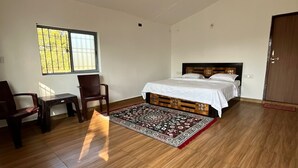 A bedroom with a wooden floor, a bed with a white bedspread, a ceiling fan, a window with bars, and a door. - Home in the Jungle (Kurai)