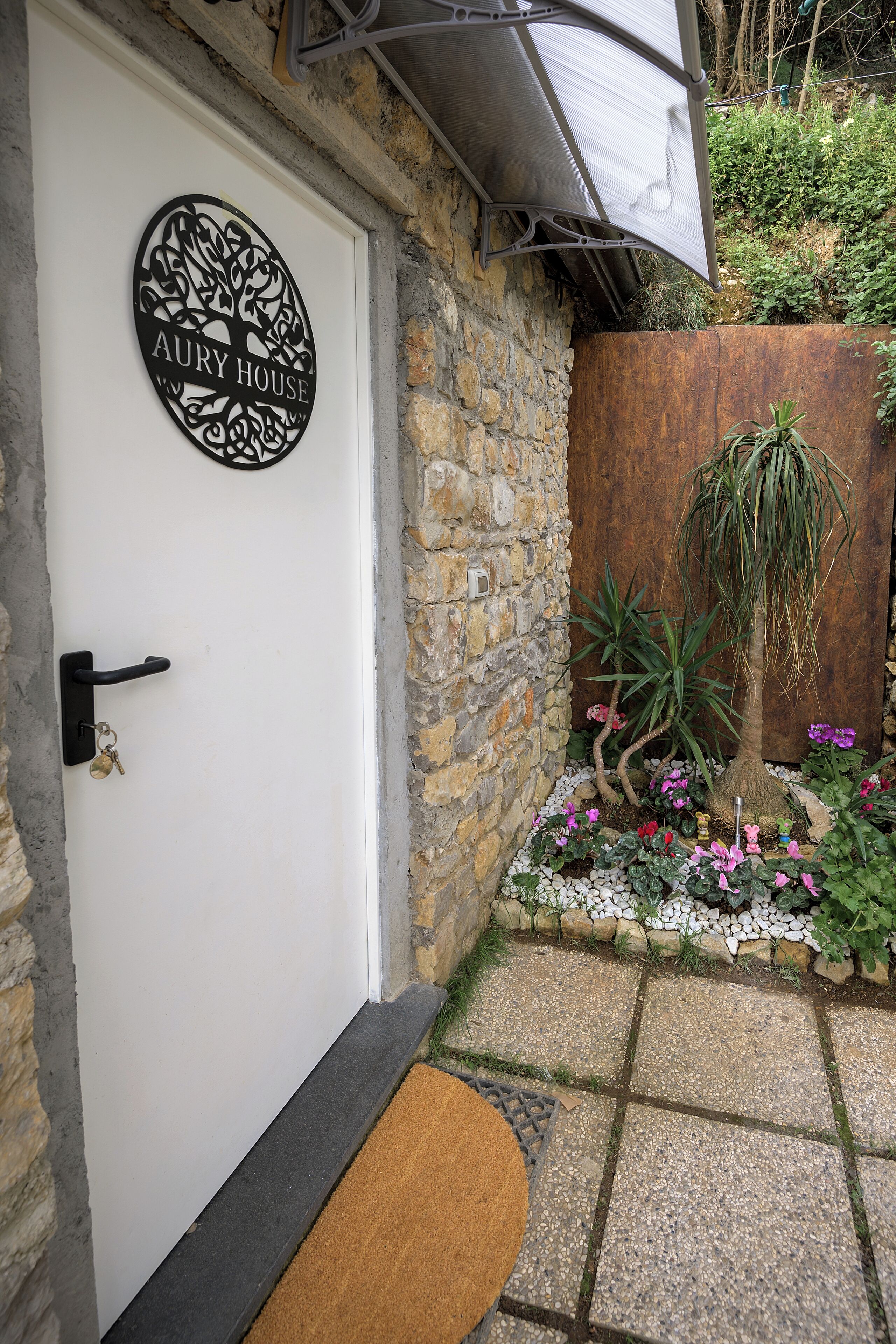 A white door with a black sign reading "AURY HOUSE" and a small garden with plants and pebbles.