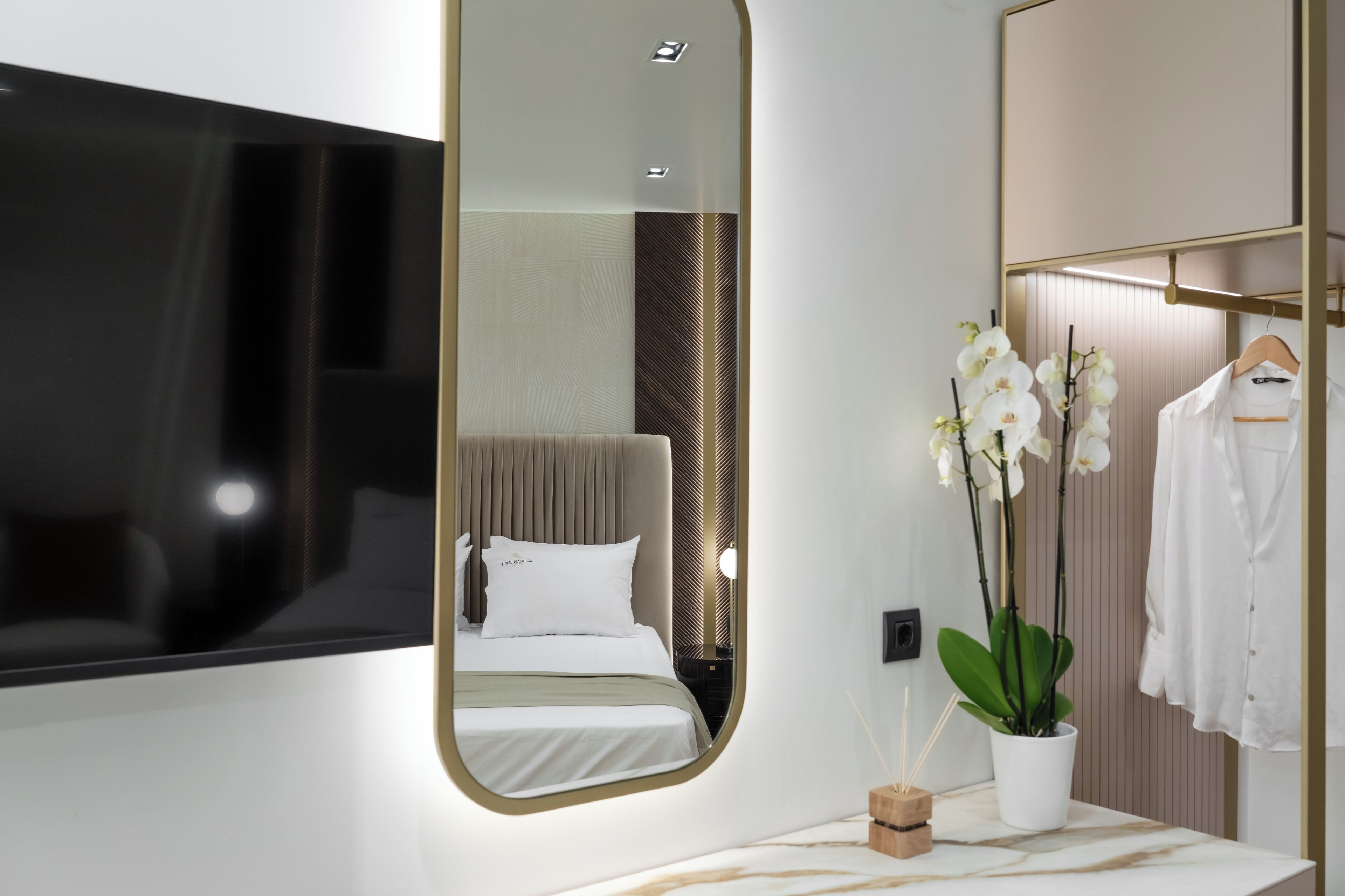 Ein modernes Hotelzimmer mit einem großen Spiegel, einem Bett mit weißen Leinen, einem an der Wand montierten Fernseher, einem Holzschrank und einer Orchideenpflanze im Topf.