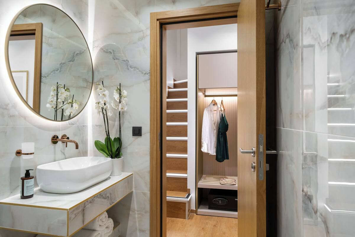 Une salle de bain moderne avec un lavabo blanc, un miroir rond et une porte en bois donnant accès à un dressing avec un portant à vêtements.