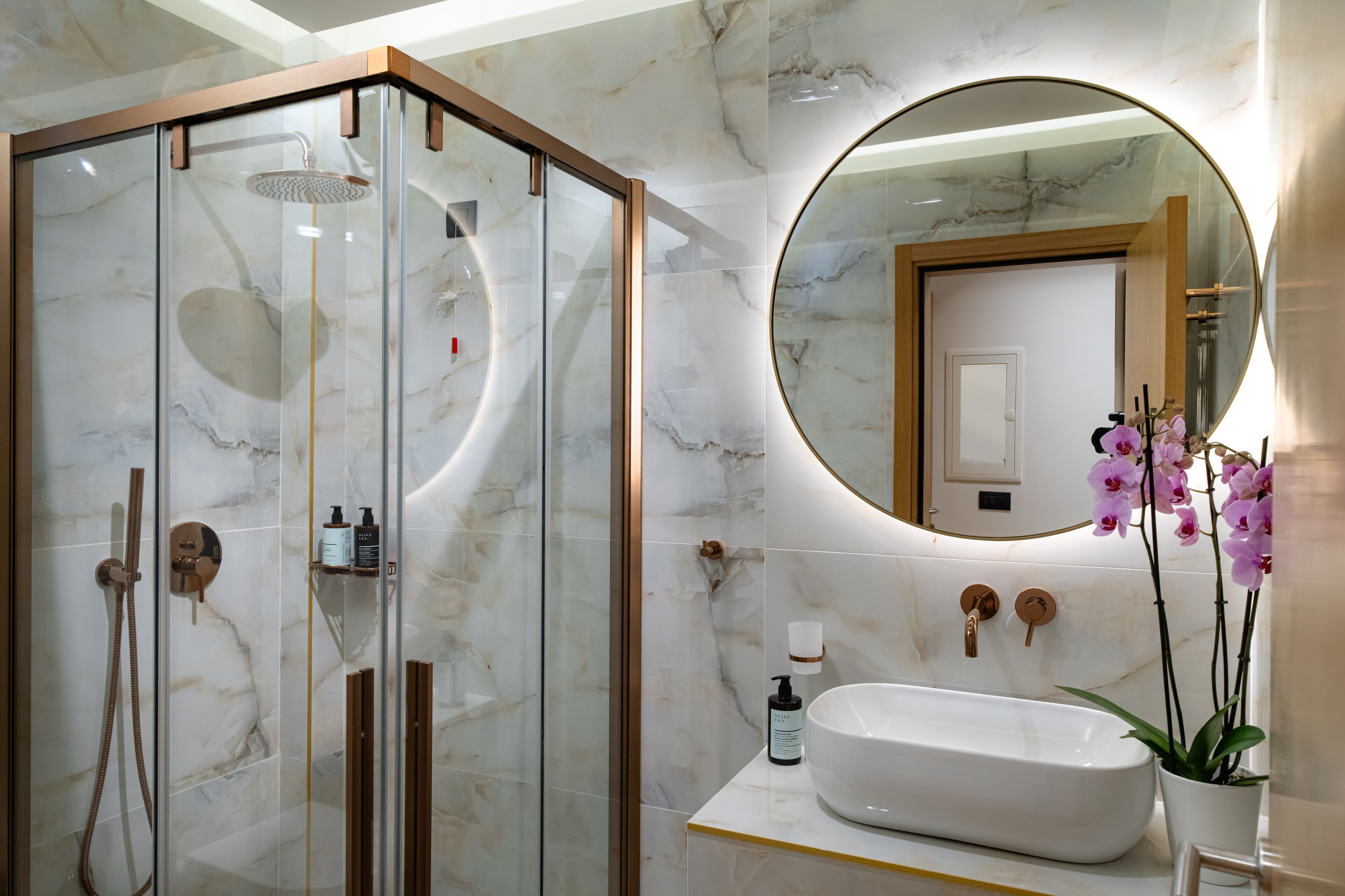 A modern bathroom with a glass-enclosed shower, a large round mirror, a white sink, and a potted orchid.