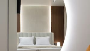 Une chambre d’hôtel moderne avec un plafond incurvé, un lit avec une literie blanche et un coin salon comprenant une chaise blanche et une petite table.