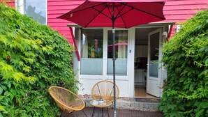 A patio area with wicker chairs and a red umbrella, surrounded by lush greenery and a pink building. - Charysma: Rosso suite | warm & stylish design | terrace | parking lot (Hameln)