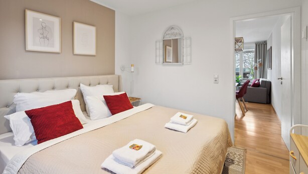 A modern bedroom with a large bed, white bedding, and red pillows. There are two framed artworks on the wall, a small mirror, and a wooden door leading to another room. - Charysma: Rosso suite | warm & stylish design | terrace | parking lot (Hameln)