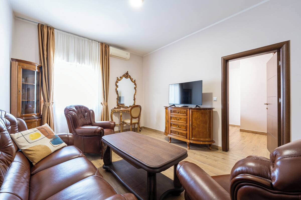 A living room with a leather sofa, a wooden coffee table, a TV on a wooden cabinet, and a large mirror on the wall.