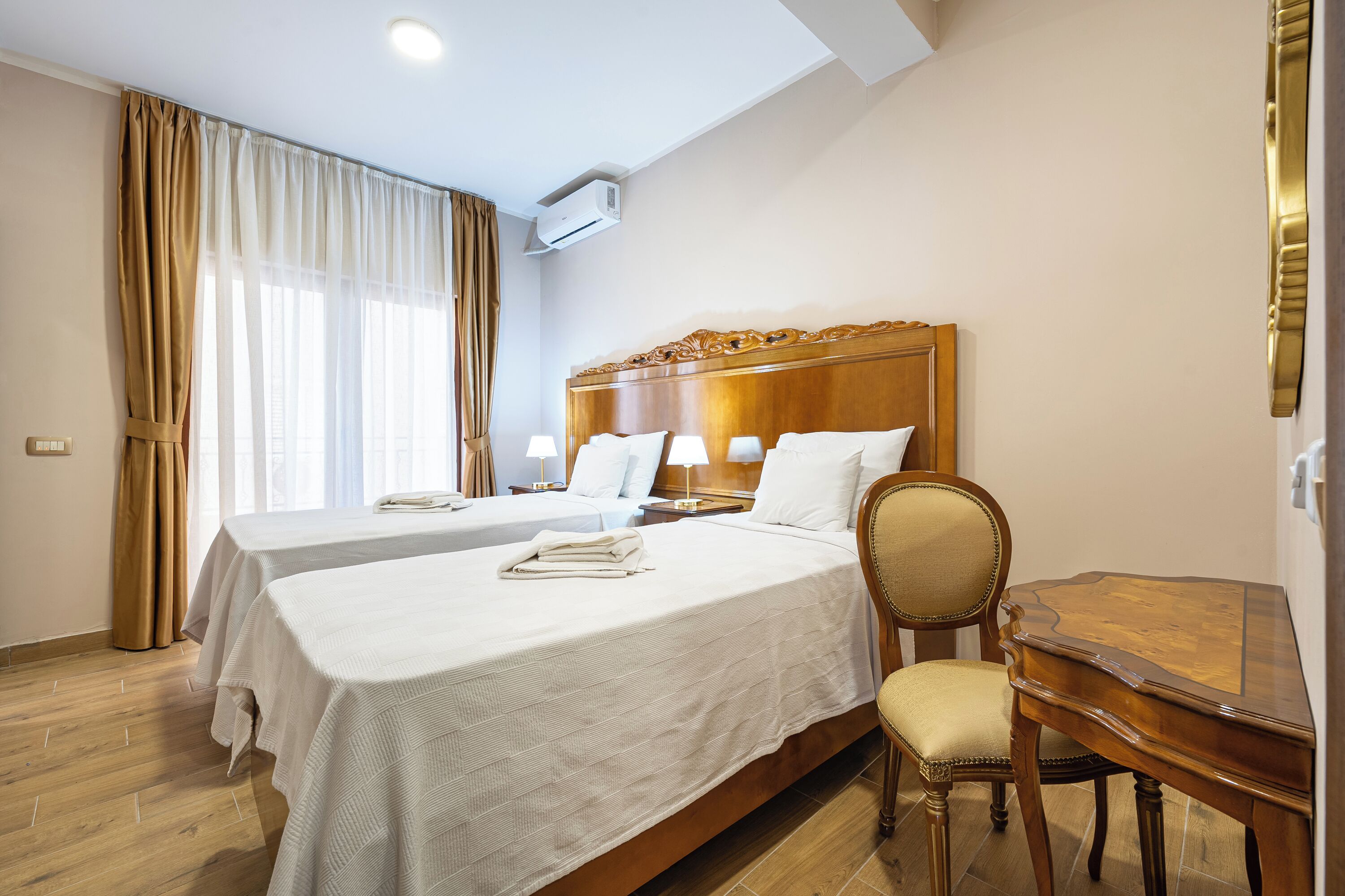 A hotel room with two beds, a wooden desk, a chair, and a window with curtains.
