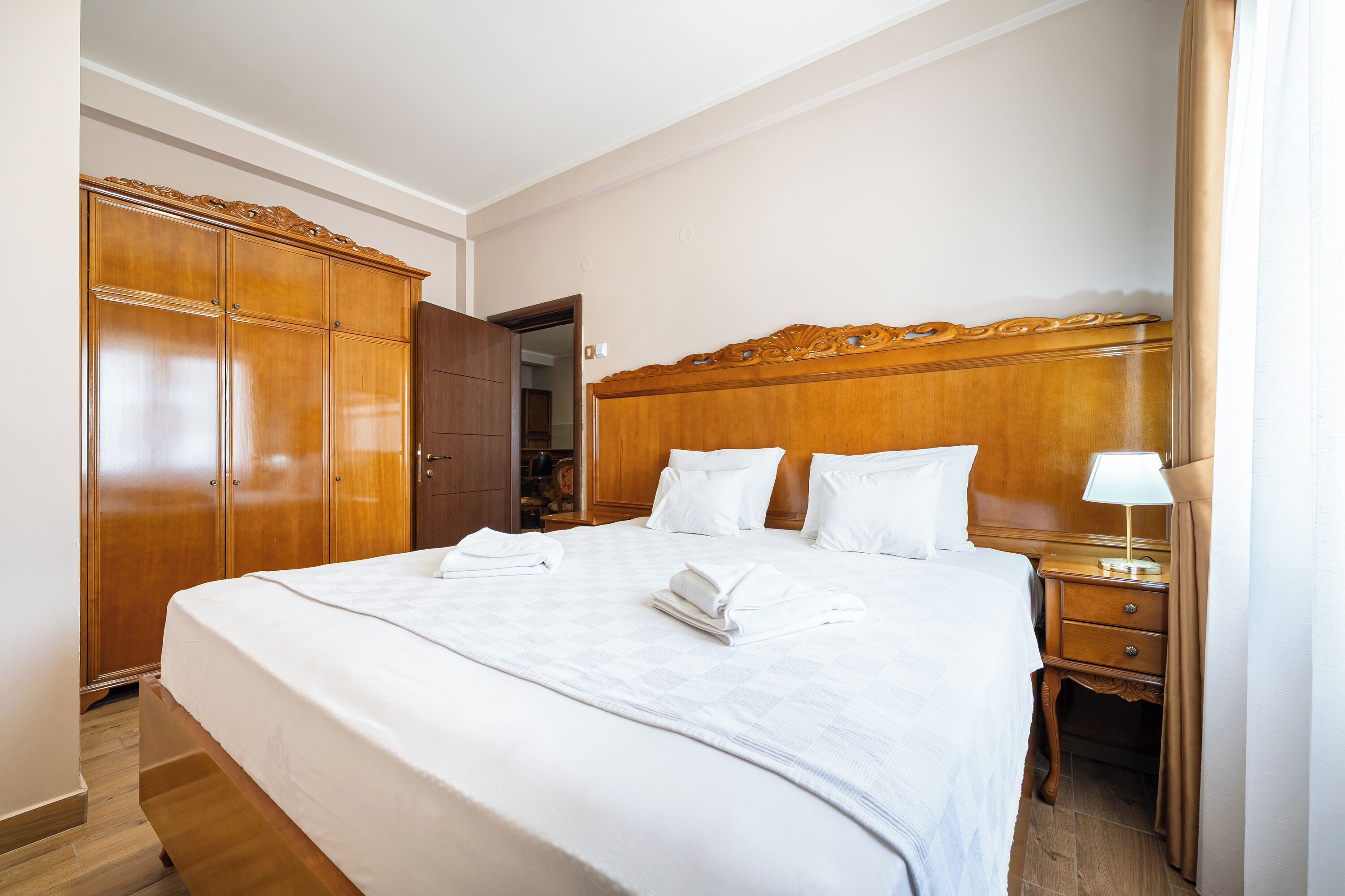 Hotelkamer met een groot bed, een houten garderobe, nachtkastje met lamp en een deur naar een andere kamer.