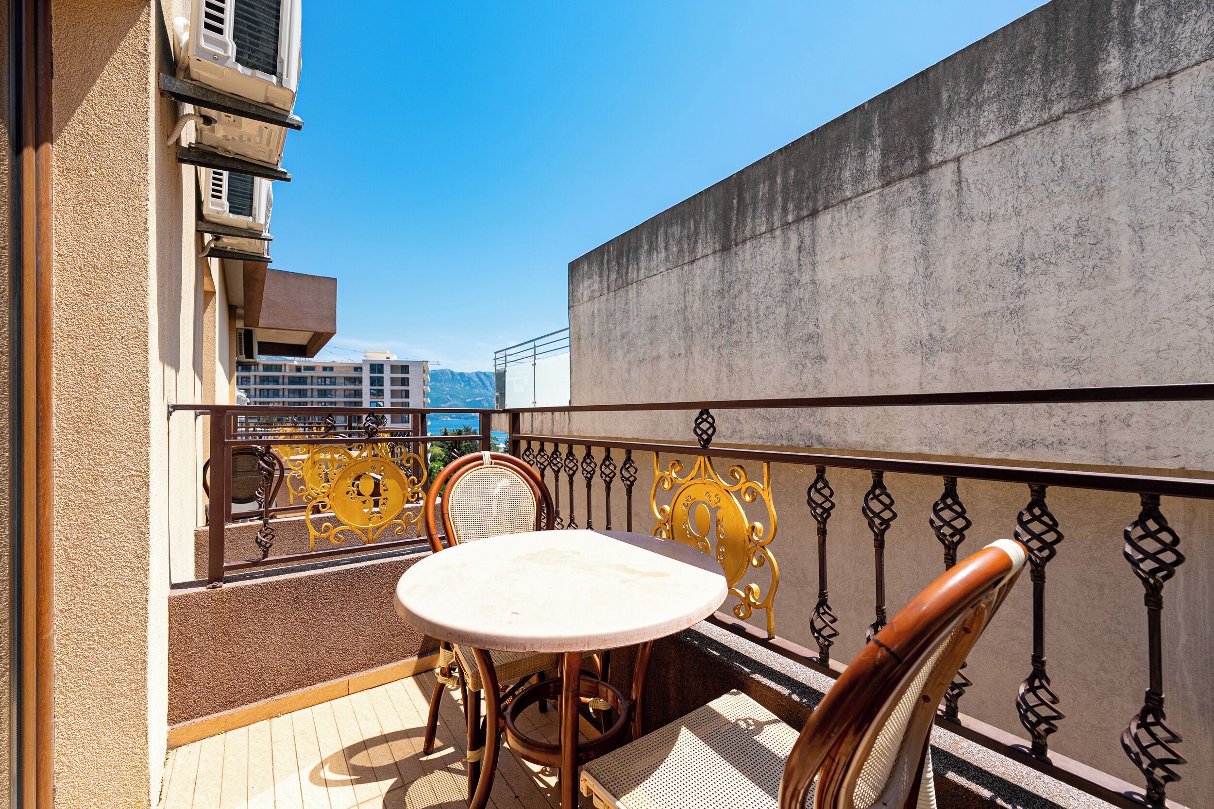 A balcony with a table and chairs, a metal railing, and a view of buildings and mountains.