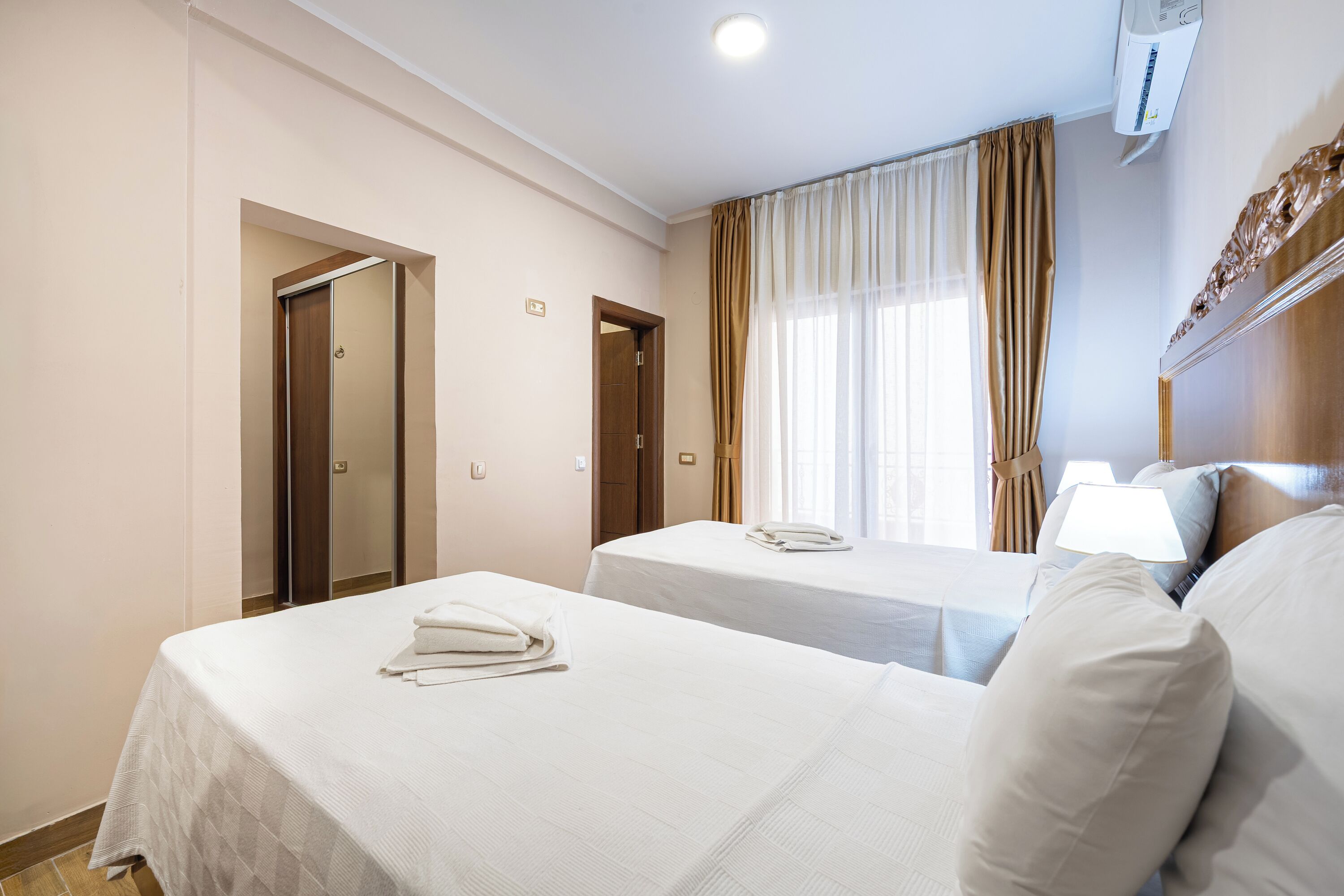 A hotel room with two beds, a window with curtains, a bedside table with a lamp, and an air conditioning unit.