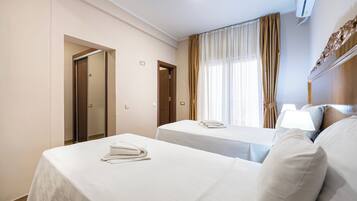 A hotel room with two beds, a window with curtains, a bedside table with a lamp, and an air conditioning unit.