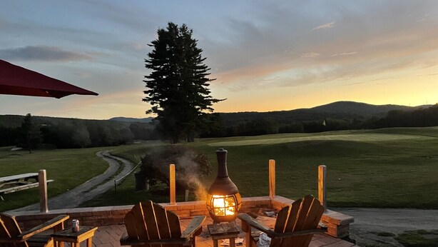 An outdoor seating area with a table and chairs, a wood-burning stove, and a view of a golf course at sunset. - New spacious-charming one bedroom cottage with loft at Wilson Lake Country Club (Wilton)