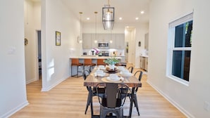 A spacious dining area with a wooden table set for six, surrounded by chairs. A kitchen with modern appliances and a large window is visible in the background. - Stylish 3BR 2BA Urban Retreat Near Lively Downtown (San Antonio)