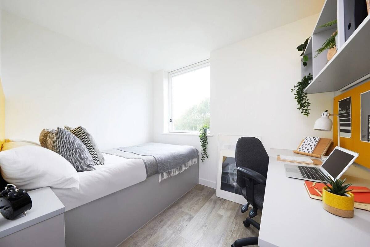 Une chambre à coucher de taille réduite, comprenant un lit, un bureau, une chaise et une fenêtre.