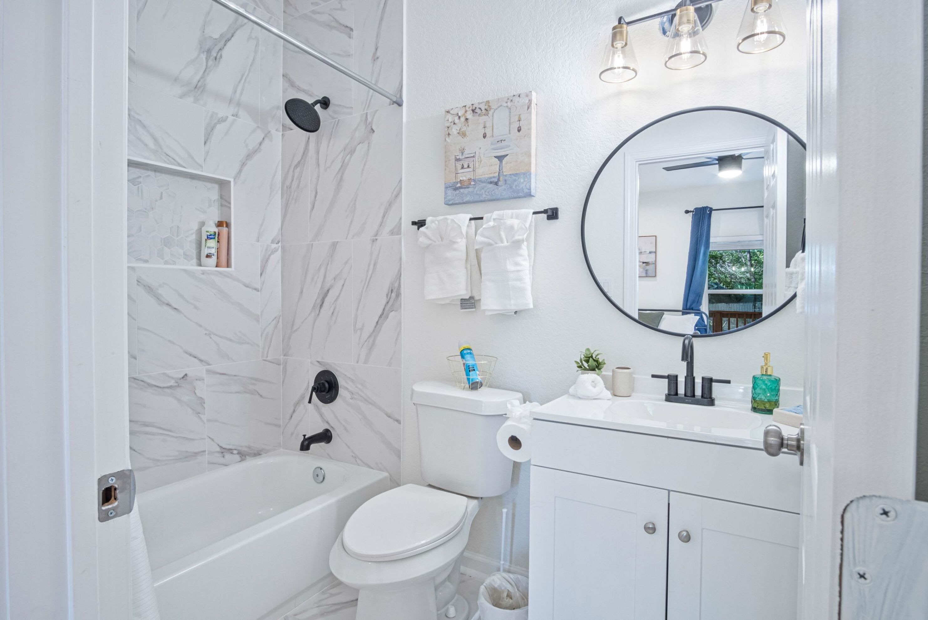 A modern bathroom with a bathtub, toilet, and sink. There is a shower curtain, a mirror, and a shelf with toiletries.