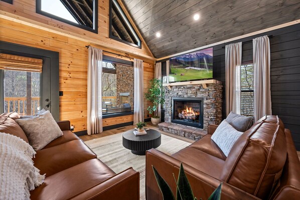 A cozy living room with a stone fireplace, leather sofas, and a large TV screen.