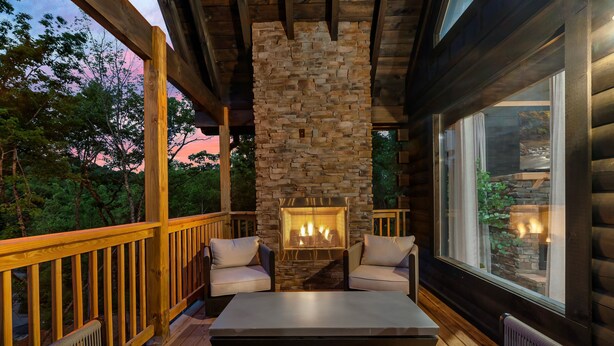A cozy outdoor seating area with a stone fireplace, wooden deck, and a view of the surrounding trees.