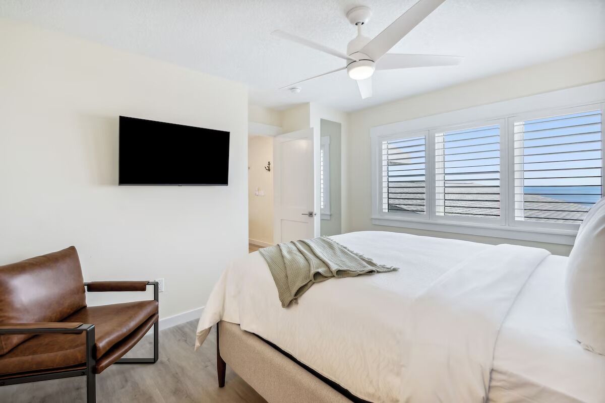 A bedroom with a bed, a chair, a ceiling fan, a TV, and a window with blinds.