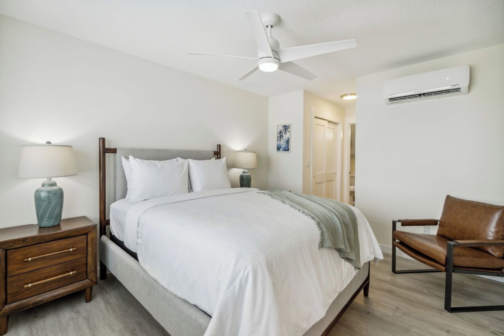 A neatly arranged bedroom with a bed, bedside tables, a chair, a ceiling fan, and an air conditioning unit.